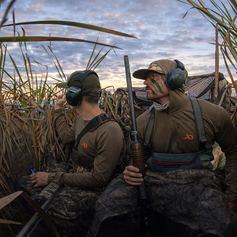 Two hunters in camouflage gear standing in tall grass with hunting rifles.